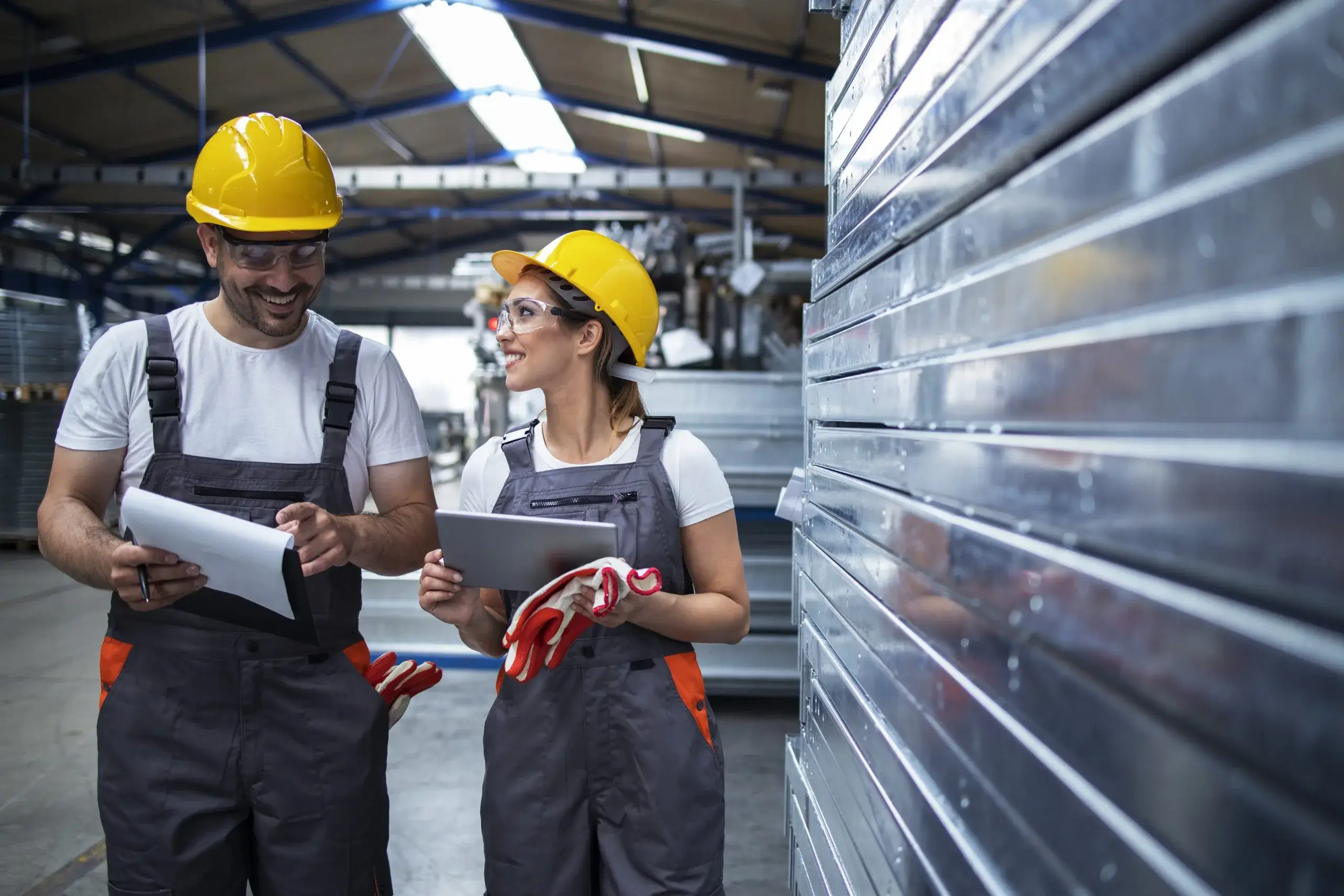 Two workers in hard hats discuss plans while holding paperwork in a warehouse