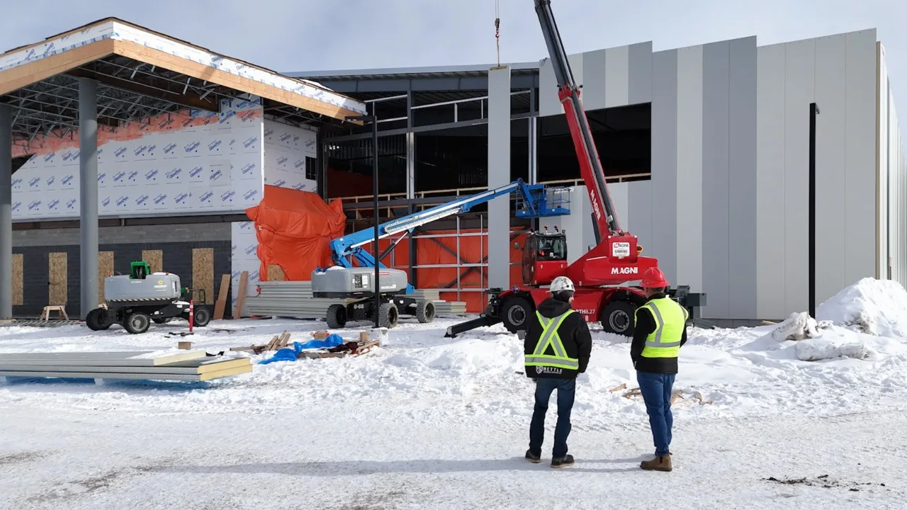 Two workers in safety vests observe construction equipment while snow covers the ground